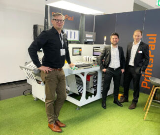 Paul Arndt, PrintsPaul; Christian Kröger, BST und Christian Dotzler, Nyquist Systems, am PrintsPaul-Stand auf der ICE Europe. Im Hintergrund die Konfektioniermaschine PPVSA420 mit einem TubeScan 100%-Inspektionssystem mit 4k-Kamera (Quelle: PrintsPaul)